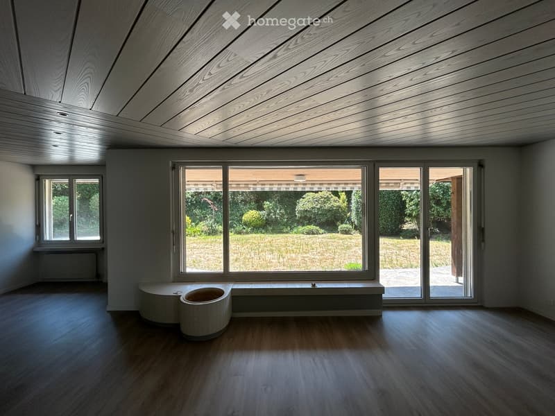 Empty room with wooden flooring, large windows, and a built-in bench along one wall. The ceiling features a sleek, modern wooden design.