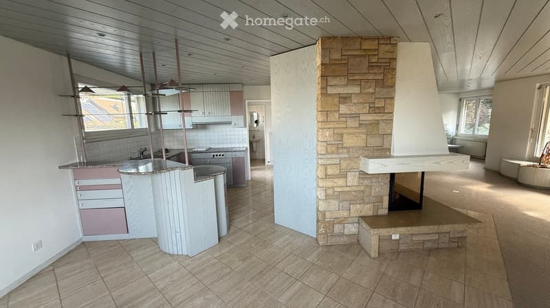 Open kitchen with central island, stone fireplace, living room, two doors to other rooms, windows, tiled floor.