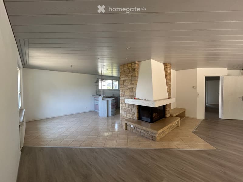 Open-plan living and dining area with a central stone fireplace, wooden flooring, and a modern kitchen area with cabinets and an island.