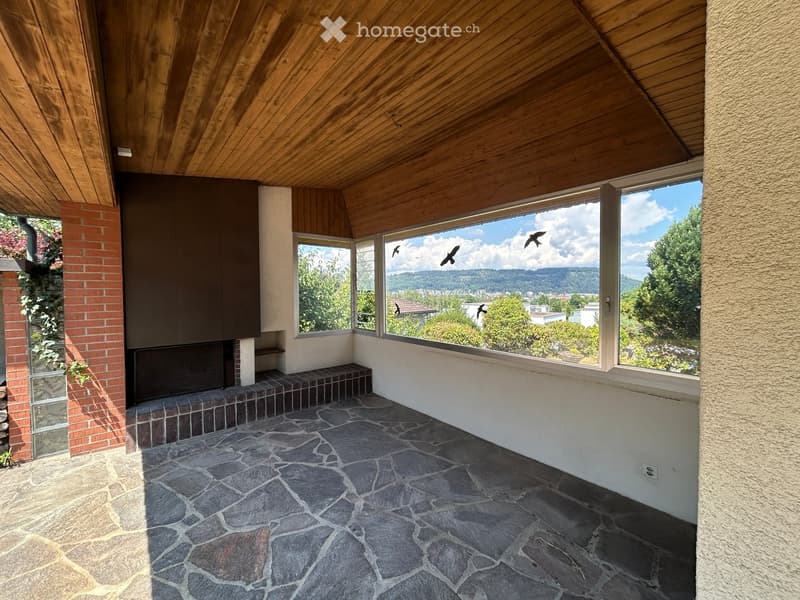 An unfurnished room with a stone-tiled floor, large windows offering a scenic view, and a fireplace area with a brick surround.