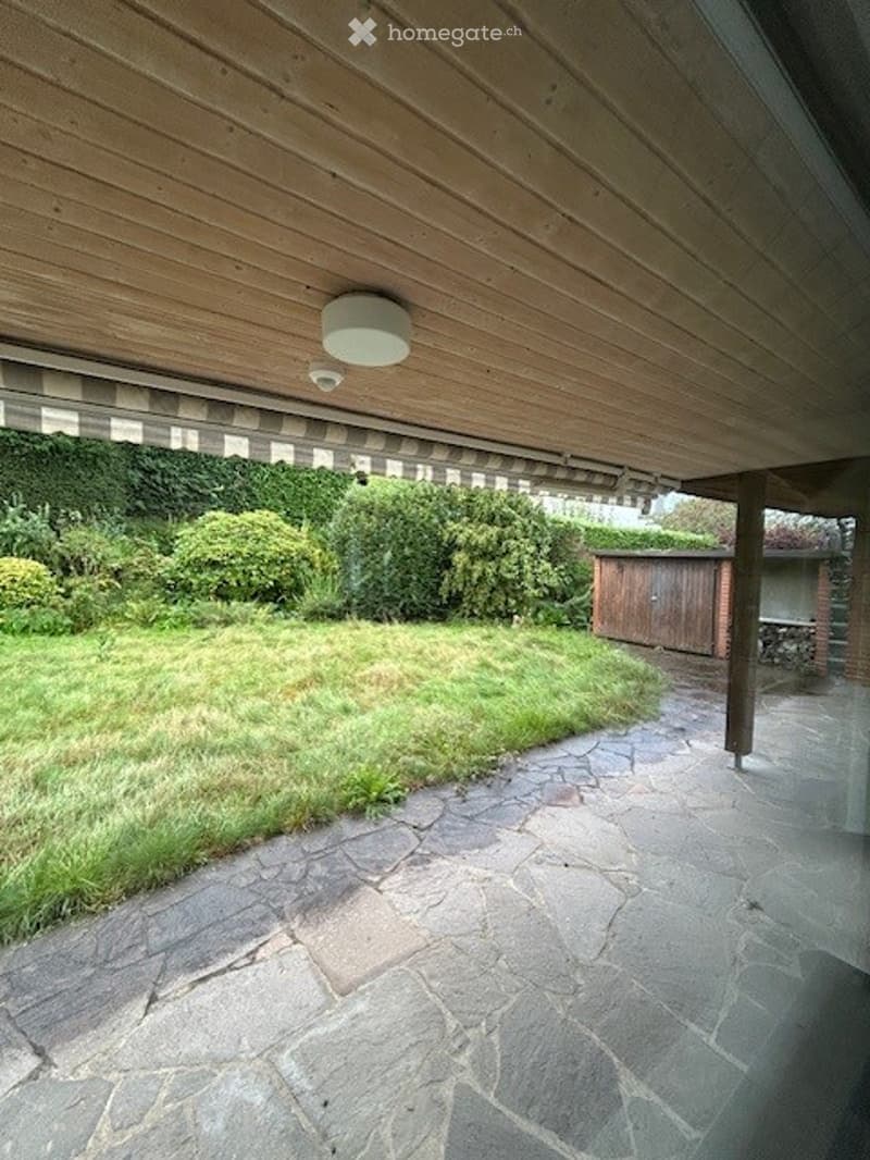 stone paved outdoor area, covered ceiling, green grass, wooden house, bushes