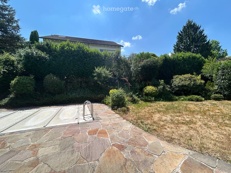 Garden area with stone-paved pathway, grassy lawn, and swimming pool, surrounded by trees and shrubs.
