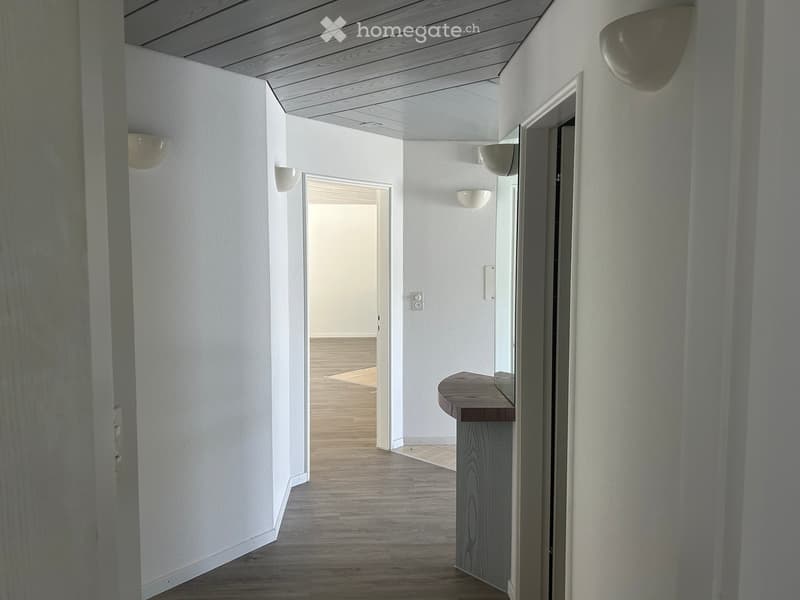 A modern hallway with light wooden flooring, white walls, and recessed lighting. A small wooden bench sits against the wall, and an open doorway leads to another room.