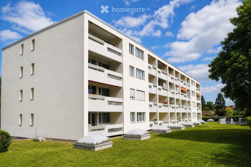 Building. A modern, white multi-story building with balconies and a flat roof, surrounded by a well-maintained lawn.