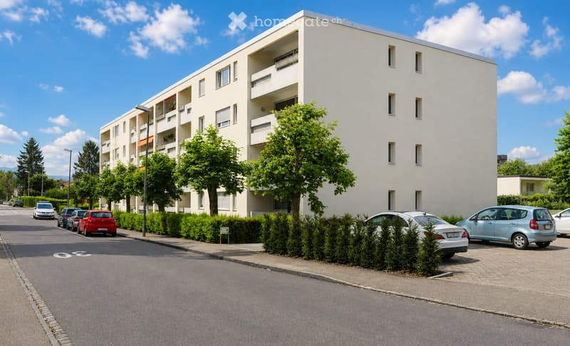 Building. Modern white apartment building with balconies, located on a street with parked cars and greenery.