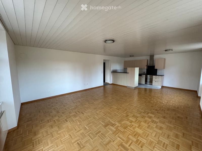 Empty room with wooden parquet flooring, white walls, and ceiling. There is a kitchen area with cabinets and appliances.