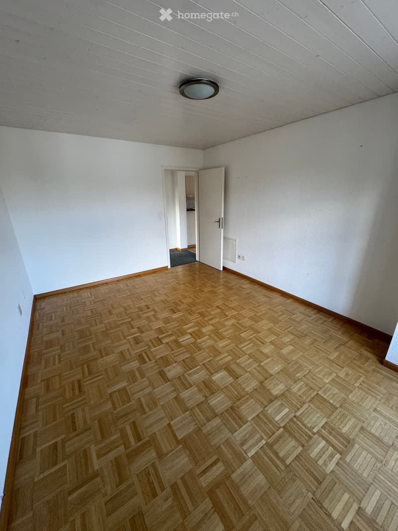 Completely empty room with white walls, wooden parquet flooring, and a ceiling light. There is an open door leading to another room.