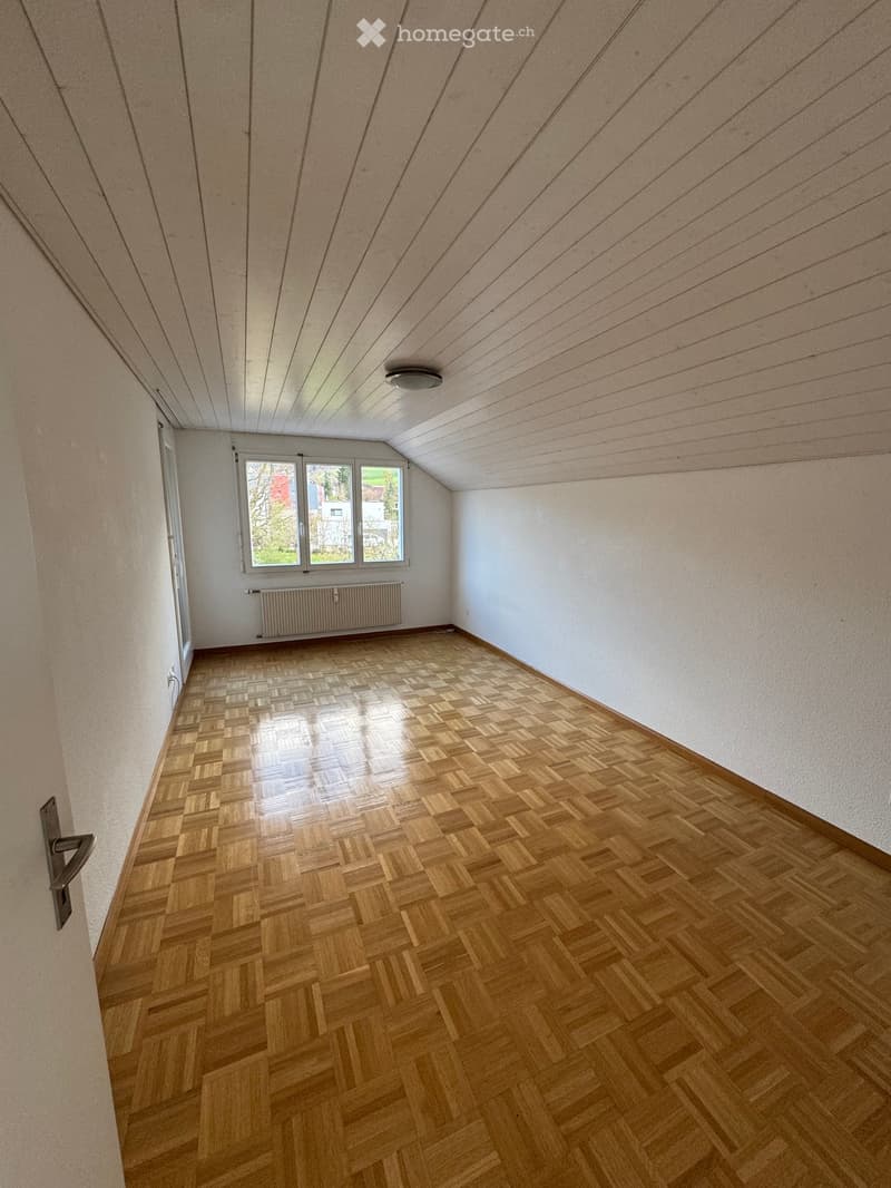 Empty room with wooden parquet flooring, white walls, and a ceiling with wooden beams. There is a window with a view of trees and buildings outside. A white radiator is mounted on the wall below the window.