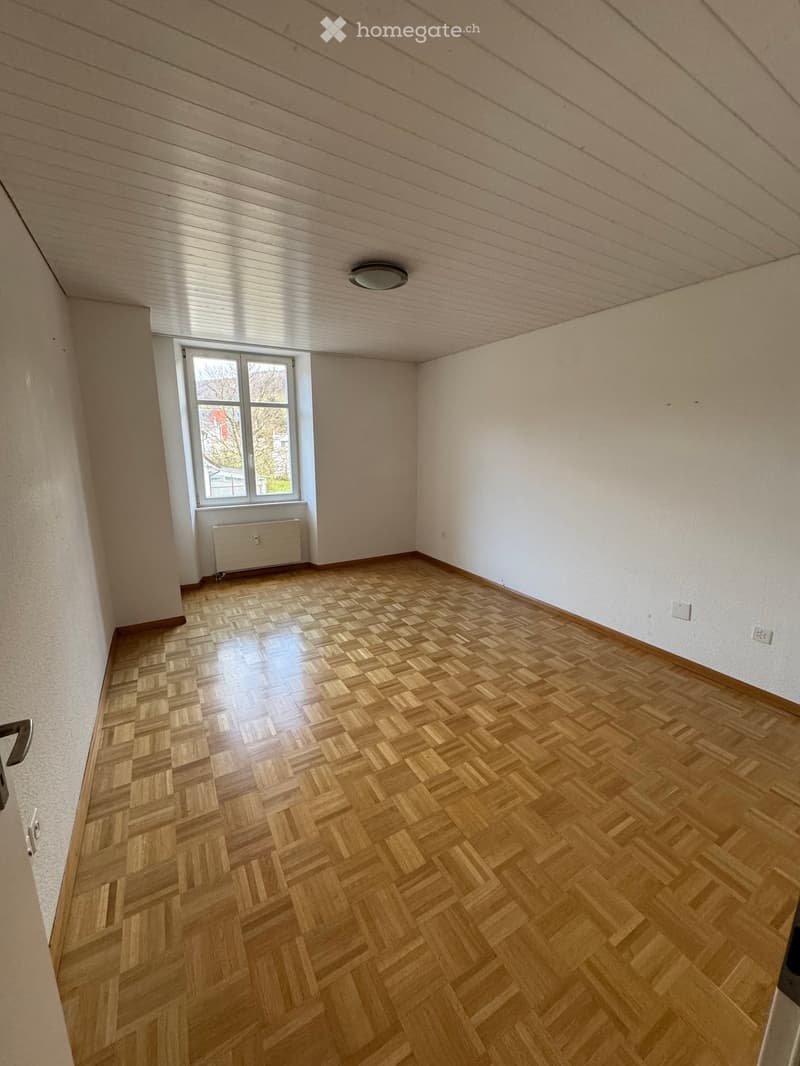 Empty room with wooden parquet flooring, white walls, and a window. There are two power outlets on the wall.