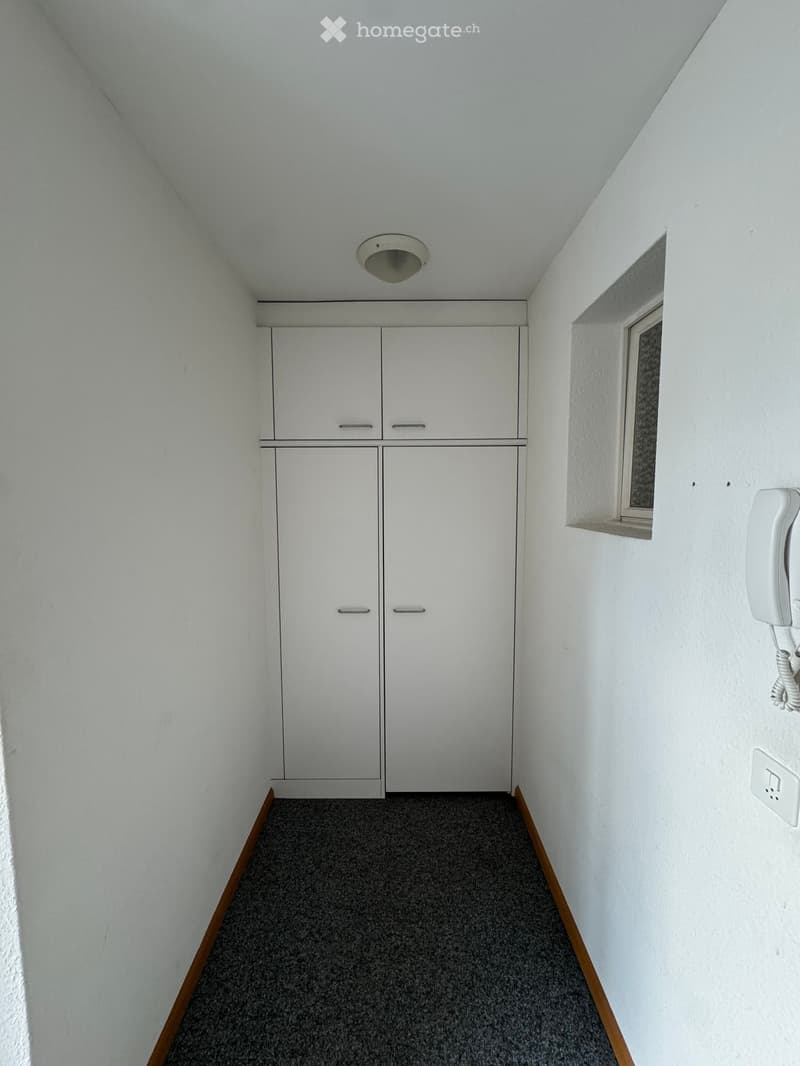 An empty room with white walls, a ceiling light, and a small window. There are two white cabinets with doors and a telephone mounted on the wall.