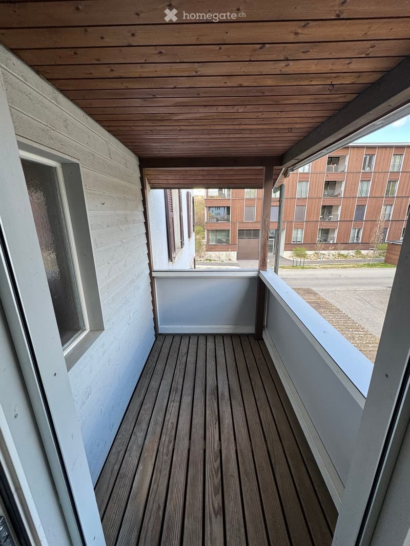 A balcony with wooden flooring and a white wall. The balcony has a white railing and a glass door. The balcony has a view of a building with glass windows and a grassy area. The balcony is empty and has no furniture.