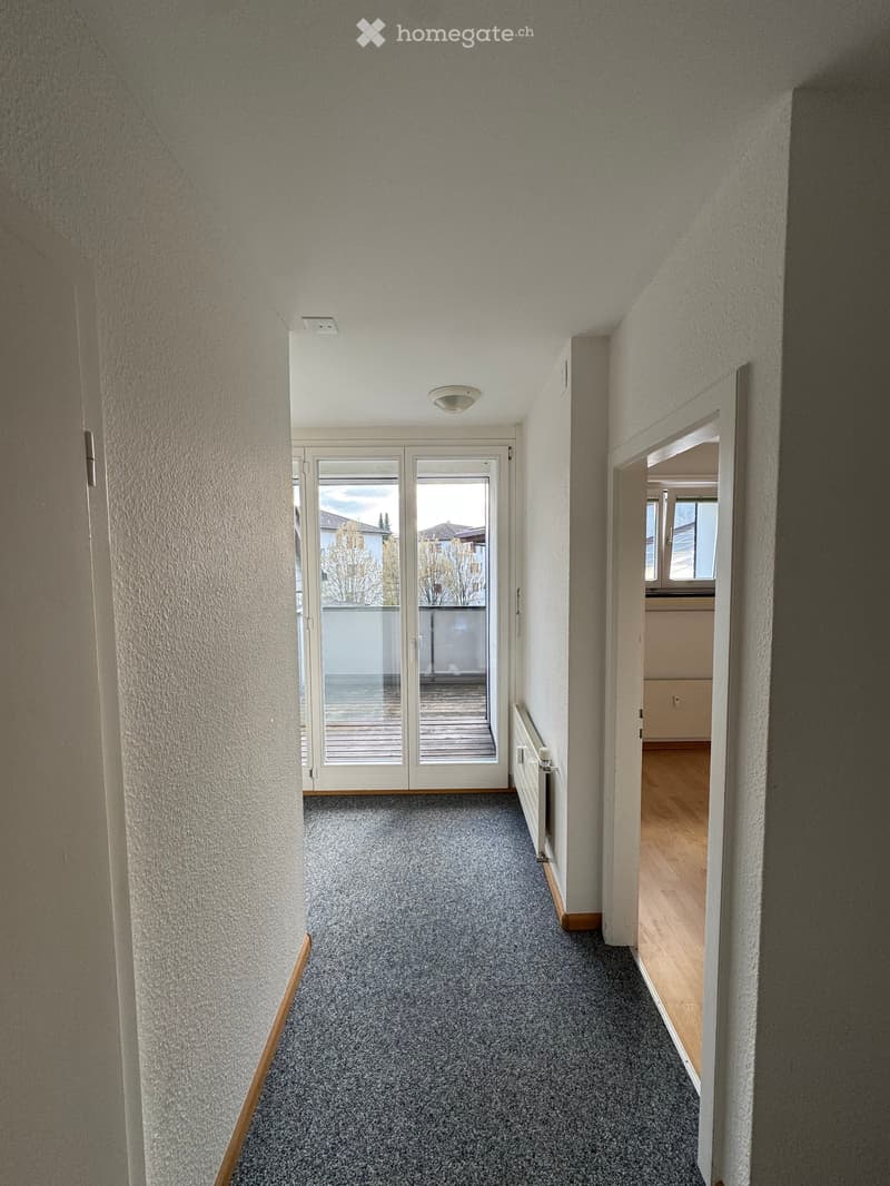 Empty corridor with white walls, gray carpet, sliding glass doors leading to a balcony, and a radiator on the wall.