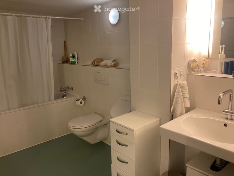 Modern bathroom with a toilet, sink, bathtub, and a mirror. The room features tiled walls, a towel hanging on a rack, and a small cabinet next to the toilet.