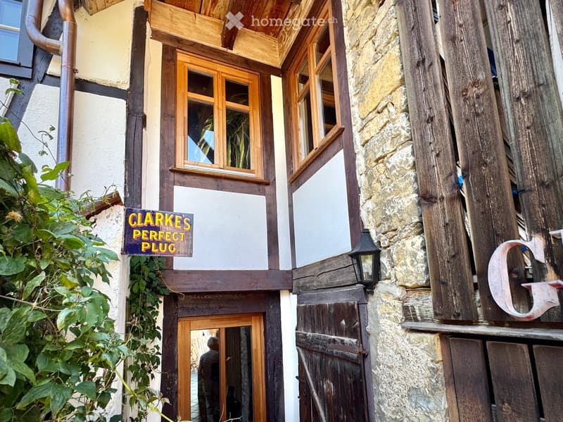 Facade of a rustic house with wooden beams, stone walls, two windows, a wooden entrance door, and a green climbing plant. It also features a sign that reads CLARKES PERFECT PLUG.