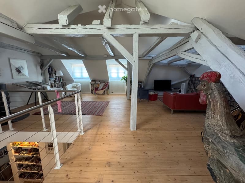 Open-plan living space with wooden floors, exposed wooden beams, large windows, and modern furniture including a red sofa, chairs, a table, and a bookshelf.
