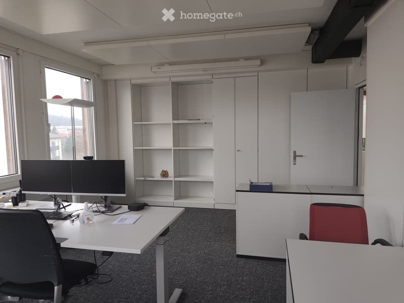 An office room with two desks, a computer on each desk, office chairs, bookshelves, white cabinets, a fluorescent lamp on the ceiling, and windows