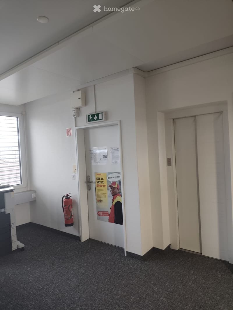 An empty room with white walls, ceiling, and floor. There is a fire extinguisher and a red fire sign on the wall. There is a window with blinds and a poster with a fire safety message on the door. There is an elevator door on the right side and a white do