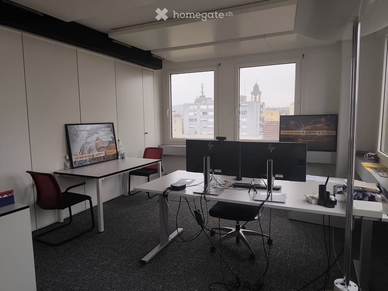 A large room with three desks, two of them are standing desks with chairs. There are large windows with an external view of buildings. The room is empty.