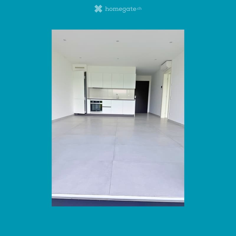 Unfurnished Room. An empty room with white walls and gray flooring. It has a kitchen area with white cabinets, a sink, and a faucet. There is a refrigerator and an oven in the kitchen area. There is a black door on the right side of the room. There are tw