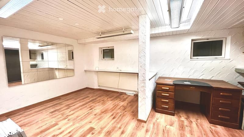 Empty room with wooden floor, white walls, large mirror, desk, and small windows.