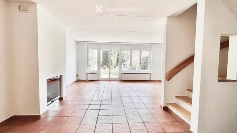 Empty room with white walls, tiled floor, and a fireplace. Large windows with sliding doors lead to a garden.