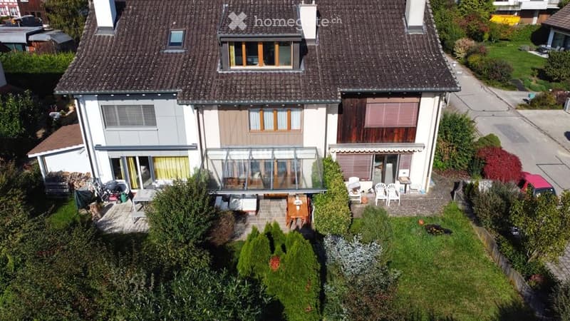 Two-story house with a brown roof, glass extension, and a garden with outdoor furniture.