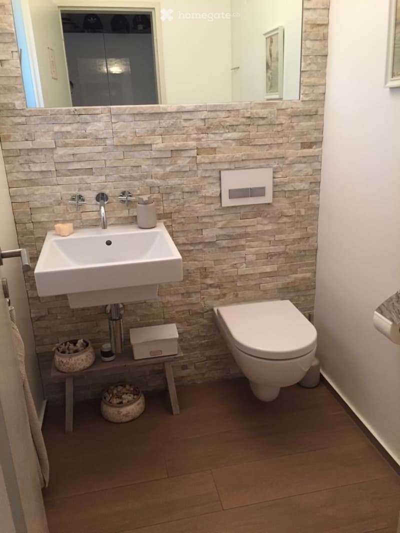 Bathroom with a white sink, faucet, and soap, a white toilet, and a mirror. The wall is made of stone tiles, and there is a small shelf with a box, bowl, and candle.