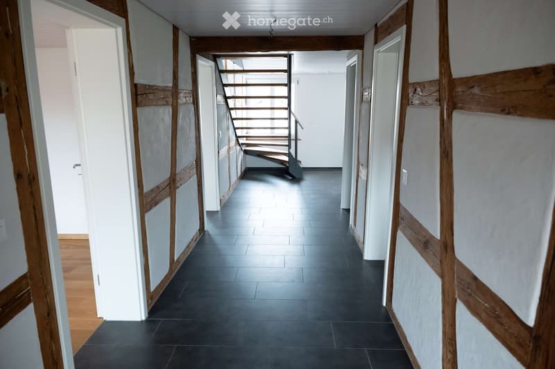 Empty corridor with black tiled floor, white walls, wooden beams, and a staircase.