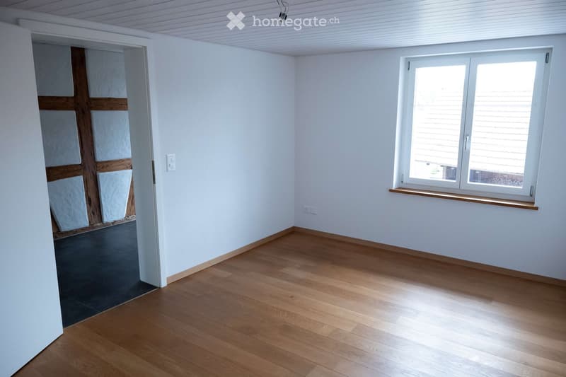 Empty room with white walls, wooden floor, and a window with a wooden sill. There is a door leading to another room with wooden beams.