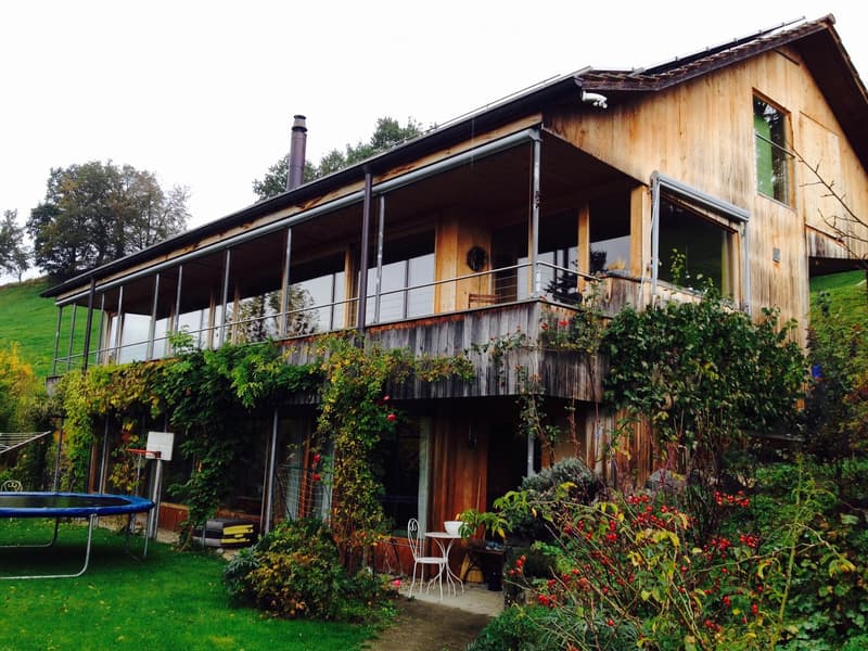 Wooden house with a balcony, surrounded by a garden with various plants and flowers.