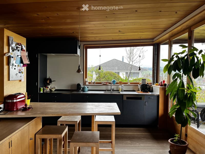 Open kitchen with wooden table, stools, and appliances. Large window with view of exterior.