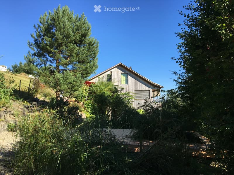 A wooden house with a red roof, nestled among trees and greenery on a hillside.