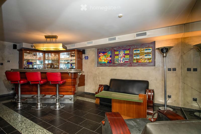 Modern bar area with red stools, a black leather sofa, a glass coffee table, and a wooden bar counter with glassware and bottles. Decorative posters adorn the wall, and a tall floor lamp stands nearby.