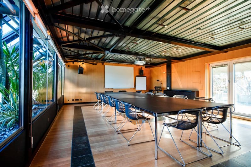 Modern conference room with a long rectangular table, multiple chairs, a large screen, and whiteboards. The room features wooden flooring, large windows with a view of greenery, and exposed metal beams on the ceiling.