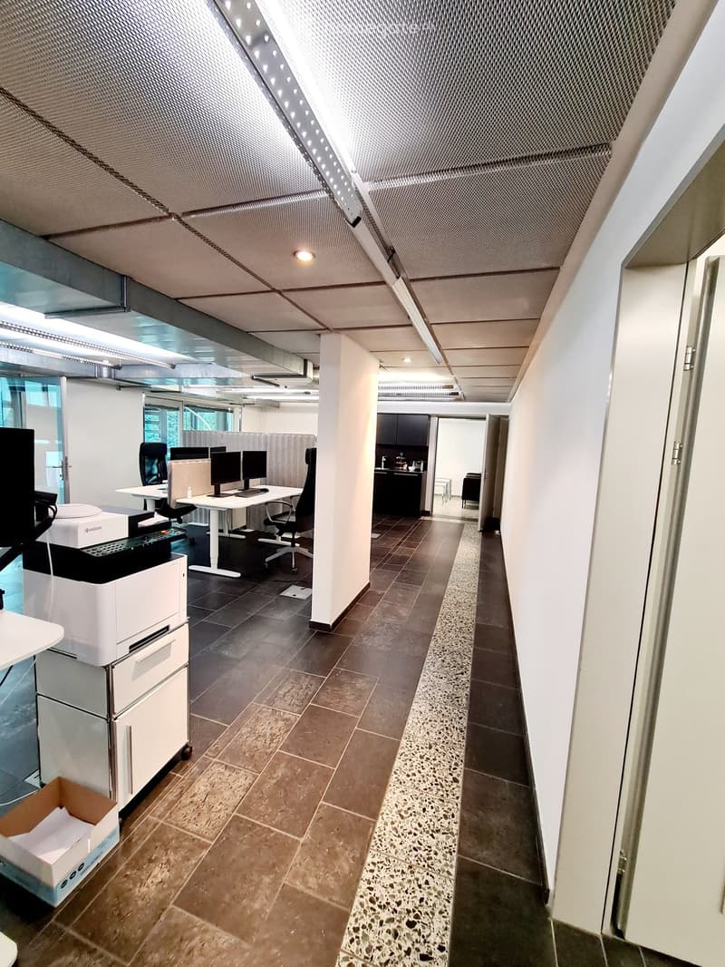 Modern office space with a desk, chairs, multiple screens, a printer, and a small kitchen area visible in the background.
