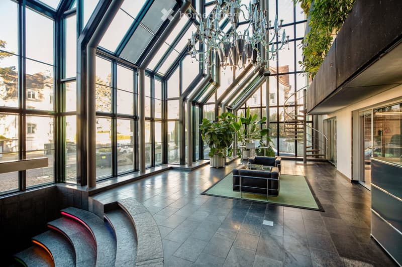 Modern, spacious lobby area with large glass windows, a chandelier, a sofa, coffee table, chairs, and potted plants.