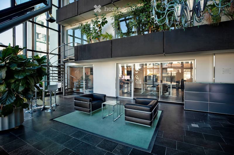 Modern, well-lit lobby area with dark tiled flooring, a green carpeted seating area, leather sofa and chairs, a glass coffee table, bar stools, and a reception desk.