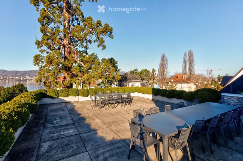 Outdoor patio with two tables and multiple chairs, surrounded by greenery and offering a scenic view of a lake and distant hills.