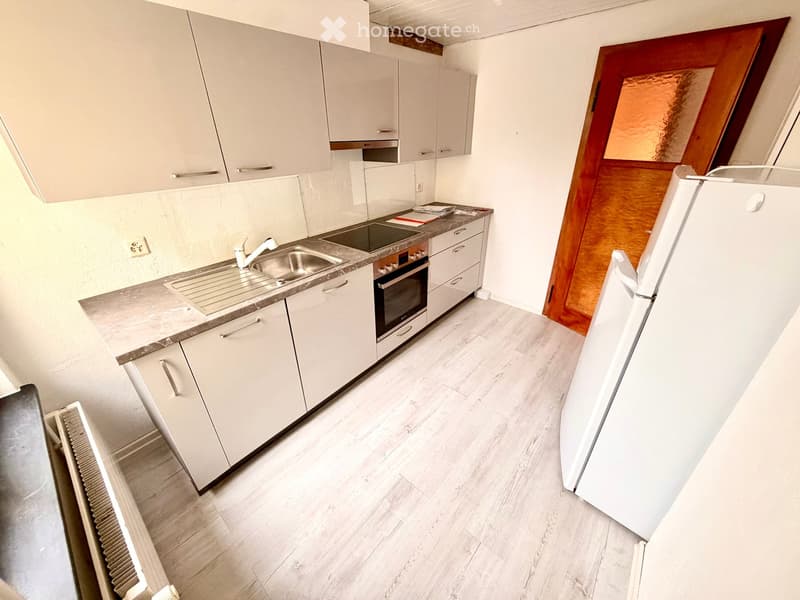 Modern kitchen with white cabinets, stainless steel sink, oven, and refrigerator. Light wood flooring and white walls.