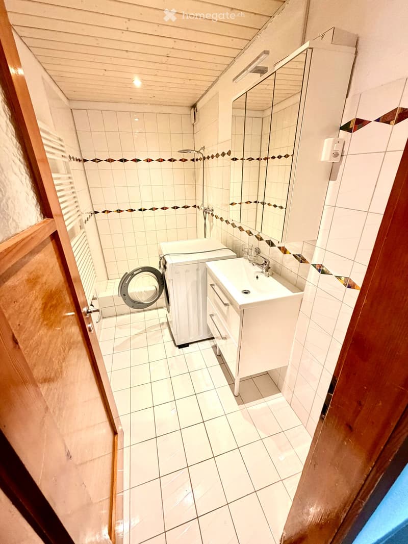 Modern bathroom with white tiles, a sink, a mirror, and a washing machine. The space features a wooden door frame and a small shower area with a glass enclosure.