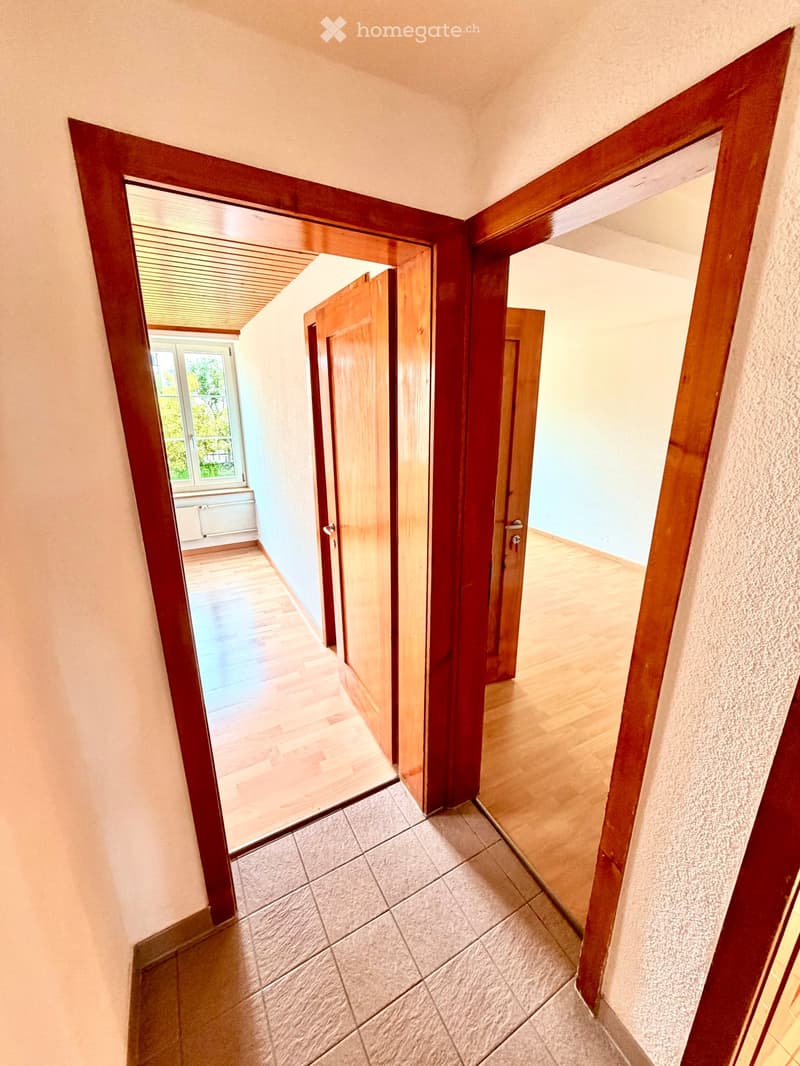 A well-lit hallway with wooden doors and a mix of tiled and wooden flooring, leading to adjacent rooms.