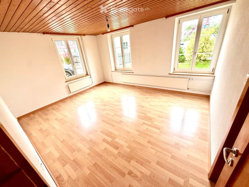 Empty room with wooden flooring, white walls, and multiple windows allowing natural light. The room features a slanted wooden ceiling and radiators under the windows.