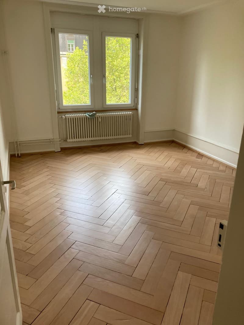 Empty room with light wooden herringbone flooring, white walls, and a radiator below a window with a view of greenery.