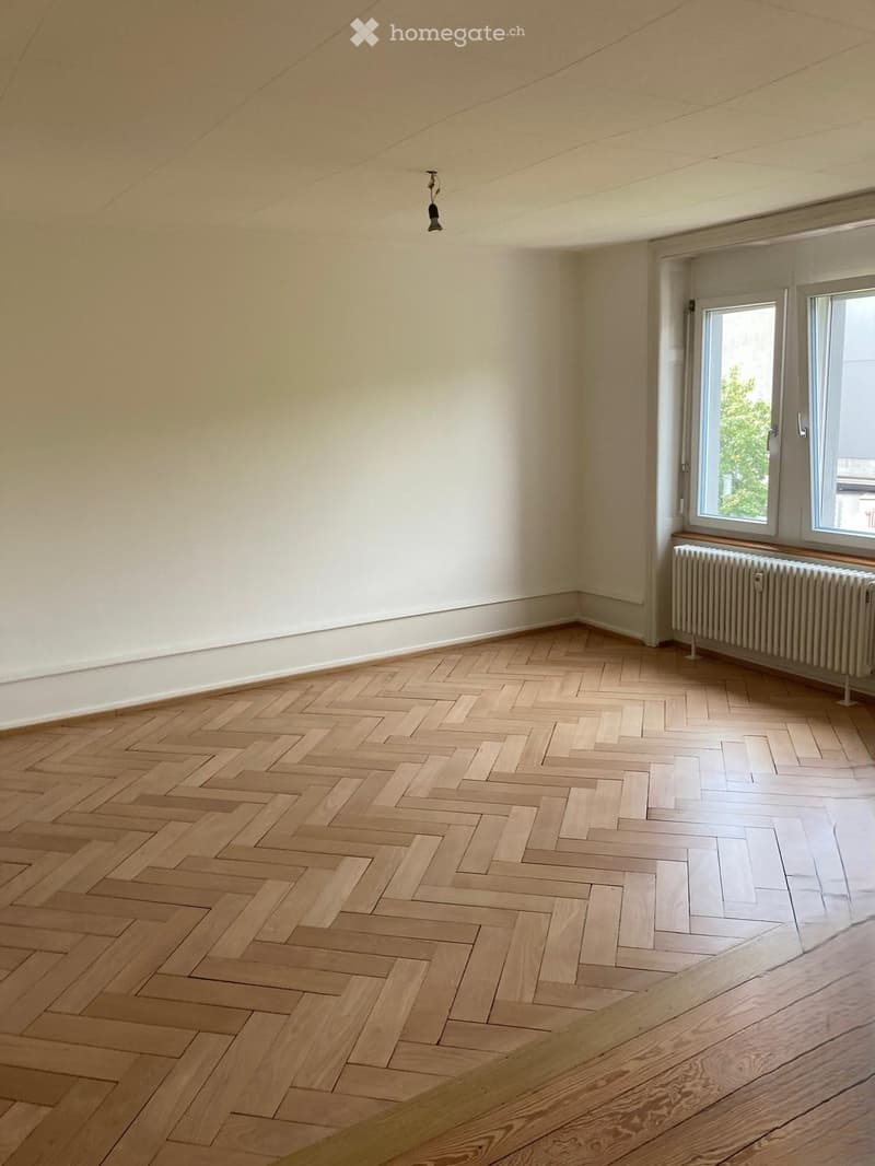 Empty room with light-colored wooden herringbone flooring, white walls, and a single window with a radiator below it.