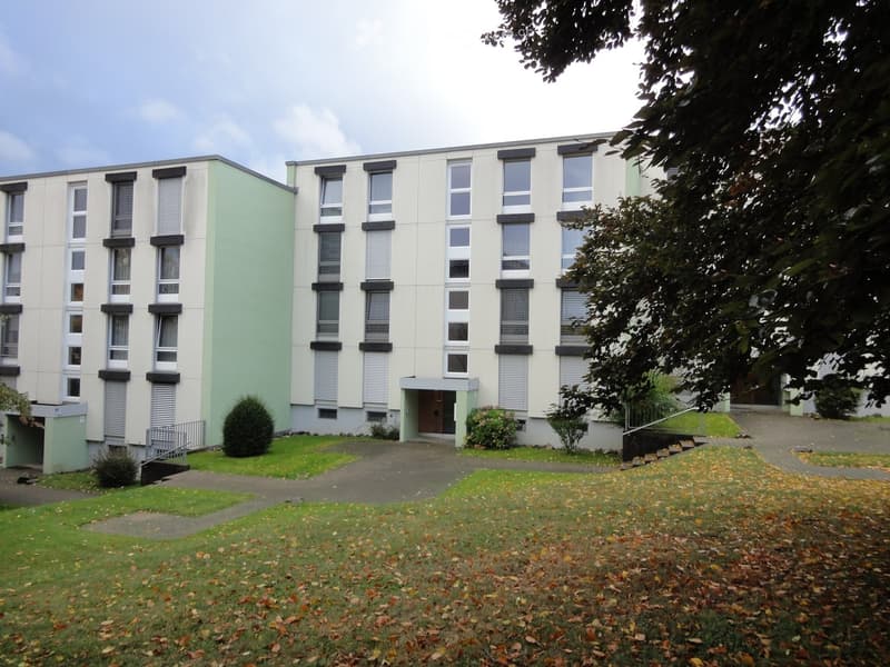 Building. A modern, two-story building with light green and white exterior walls, multiple windows with shutters, and a small entrance area. The building is surrounded by a well-maintained lawn and small shrubs.