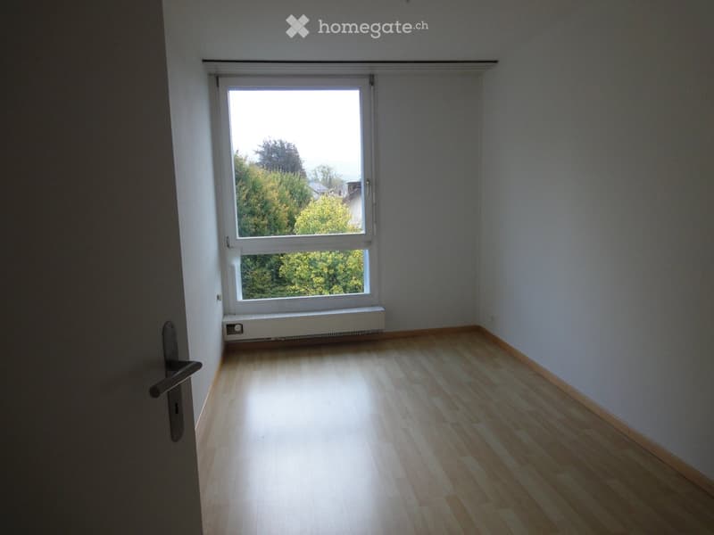 Unfurnished Room. Empty room with light wooden flooring, white walls, and a large window with a view of trees outside.