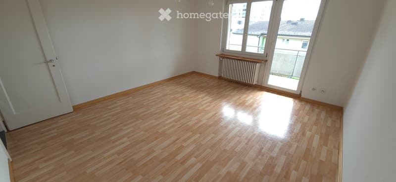 Empty room with wooden flooring, white walls, and a large window with a view of the outside. The room has a radiator and a door visible.