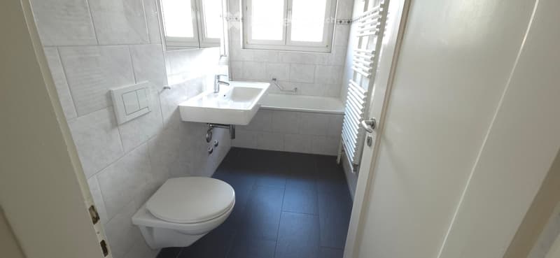 Modern bathroom with white tiles, a toilet, a sink, a bathtub, and a radiator. The space is well-lit with natural light from windows.