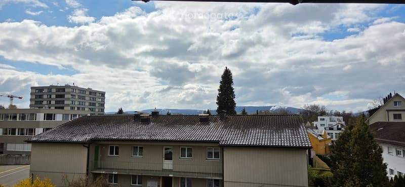 A multi-story residential building with a tiled roof, multiple windows, and a balcony, surrounded by other buildings and trees under a partly cloudy sky.