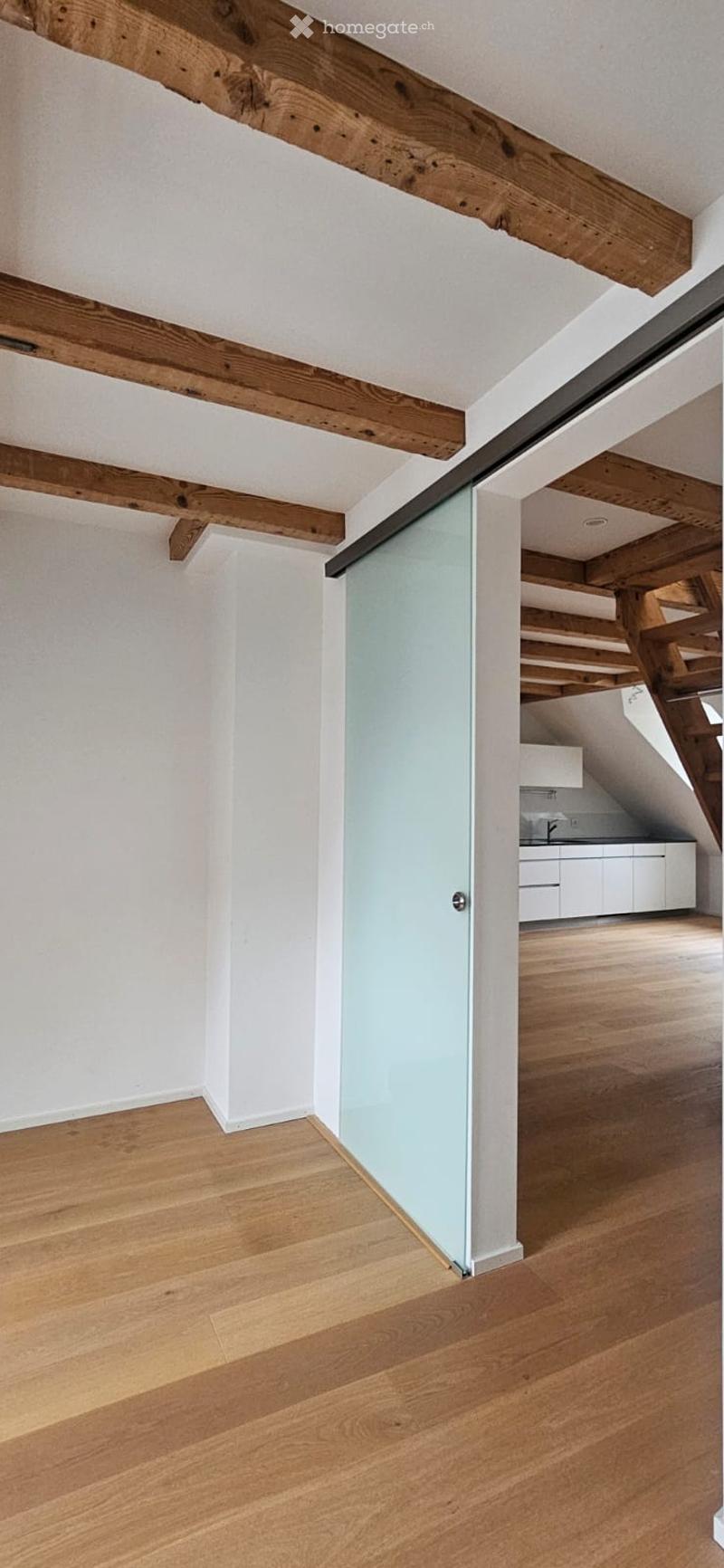 Empty room with light wooden flooring, white walls, exposed wooden beams on the ceiling, and a glass partition leading to another area.